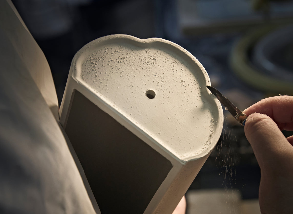 A close-up of a ceramic mug or cup being carefully trimmed with a small tool, with fine dust falling. A hand is visible on the right side, and the background is a softly blurred workshop setting.