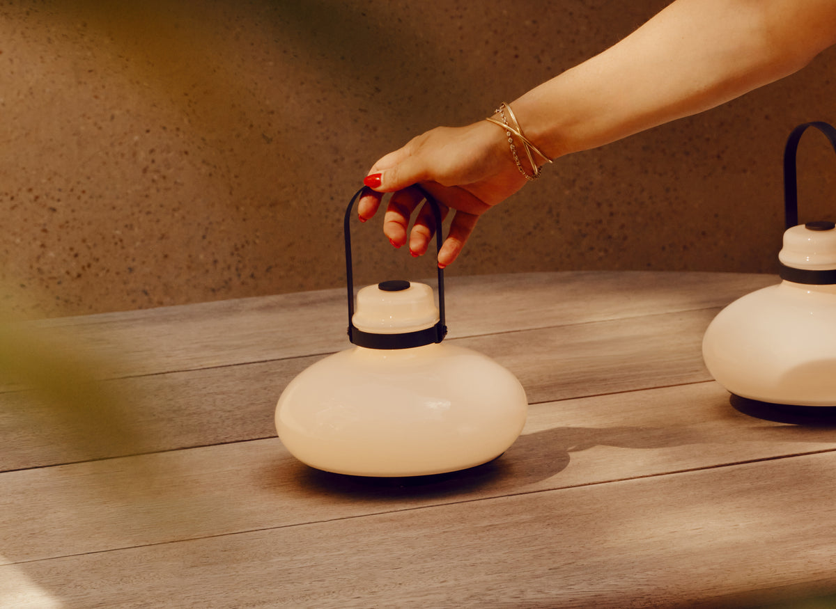 A hand with red nail polish and gold bracelets holds the handle of a modern, rounded off-white lantern with a black metal handle on a wooden table.