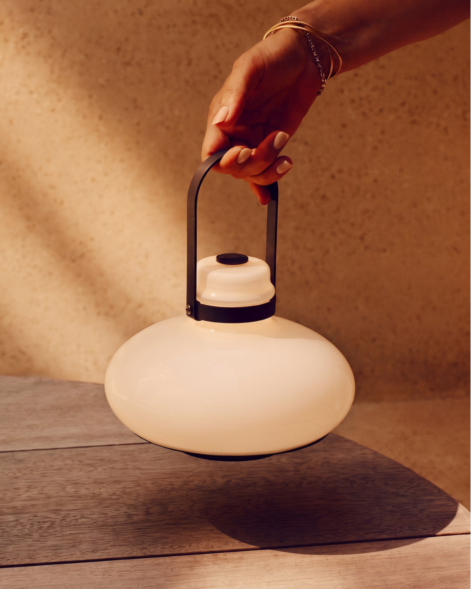A hand with gold bracelets holds the handle of a modern, rounded white lantern with a black metal handle and a warm glowing light, set against a textured beige wall and wooden table.