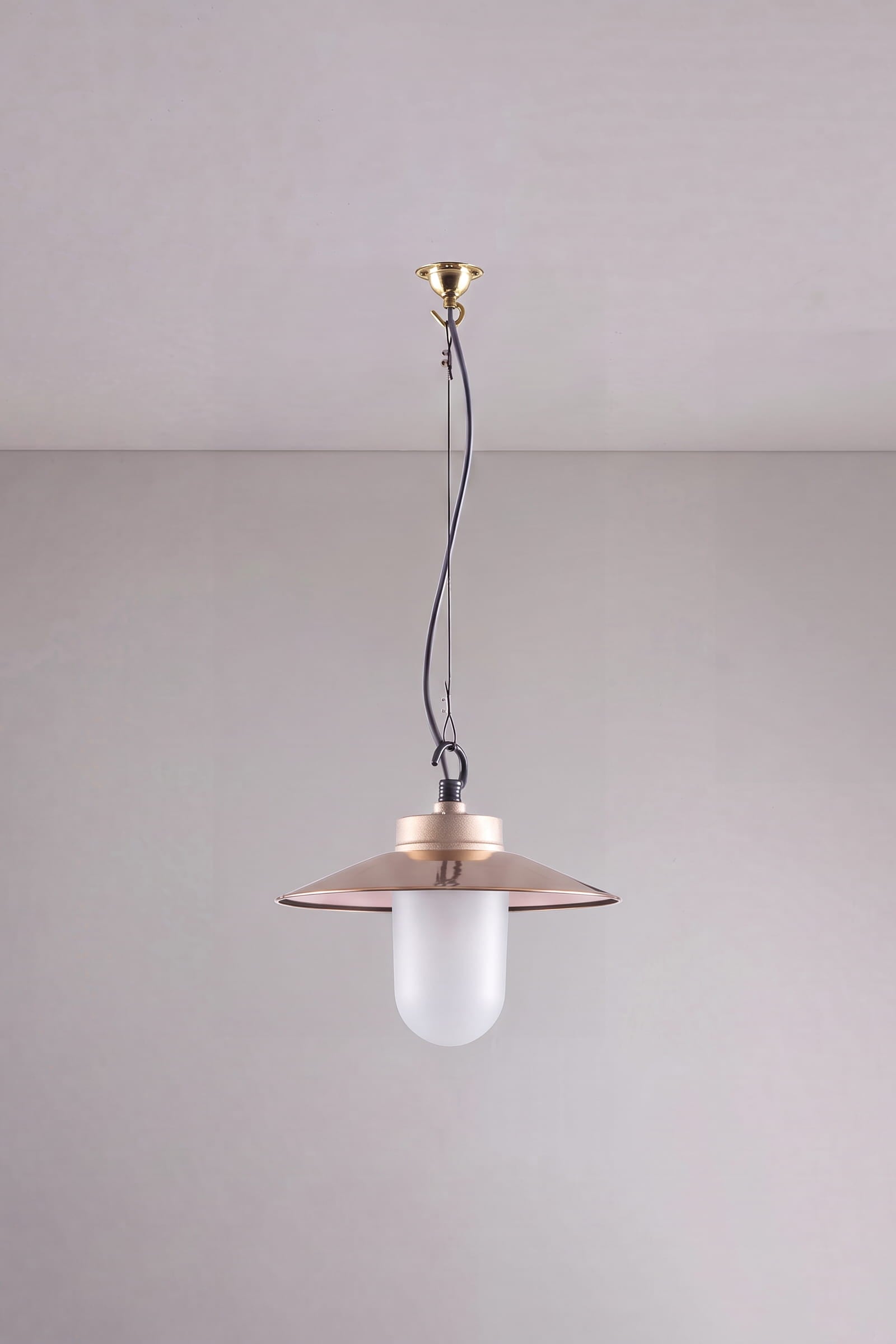 A modern pendant light with a wide, shallow bronze-colored metal shade and a frosted white glass bulb, suspended by a black cord from a brass ceiling canopy against a neutral gray wall.